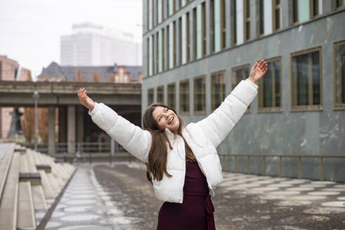 Carefree young woman with arms raised standing on footpath in city - BFRF02451
