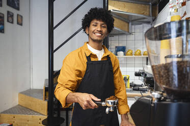 Smiling barista wearing apron and holding portafilter at coffee shop - ALKF00939