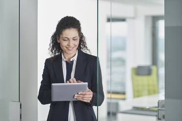 Lächelnde Geschäftsfrau, die einen Tablet-PC benutzt und in einem Büroeingang steht - RORF03676