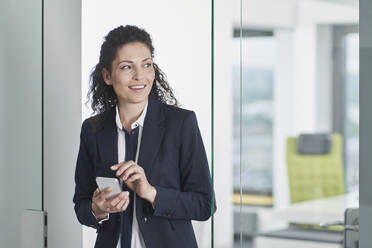 Lächelnde reife Geschäftsfrau mit Smartphone in der Bürotür stehend - RORF03680