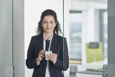 Smiling businesswoman standing with mobile phone in office doorway - RORF03682