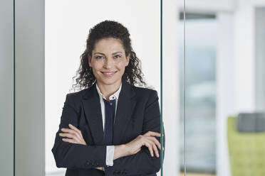 Happy businesswoman standing with arms crossed near glass door in office - RORF03688