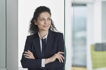 Smiling businesswoman standing with arms crossed near glass door in office - RORF03689