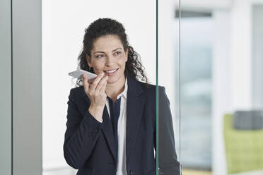 Lächelnde Geschäftsfrau, die in der Nähe einer Glastür im Büro mit einem Smartphone telefoniert - RORF03690