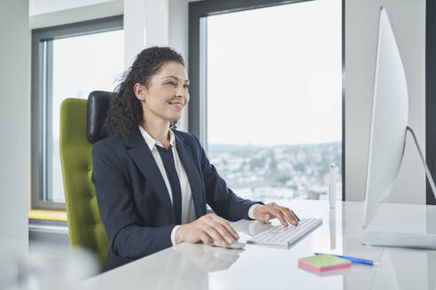 Lächelnde Geschäftsfrau arbeitet am Computer am Schreibtisch im Büro - RORF03699