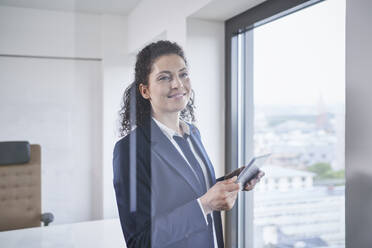 Lächelnde reife Geschäftsfrau stehend mit Tablet-PC in der Nähe eines Glasfensters - RORF03707