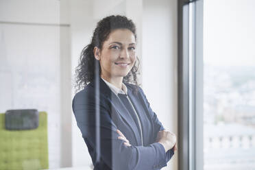 Smiling businesswoman with arms crossed seen through glass in office - RORF03713