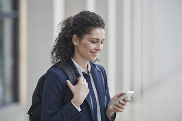 Smiling mature businesswoman with backpack using smart phone - RORF03718