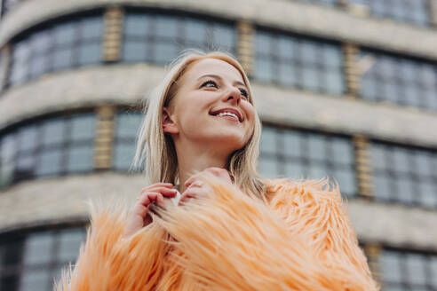 Lächelnde Frau mit pfirsichfarbenem Pelzmantel vor einem Gebäude - VSNF01572