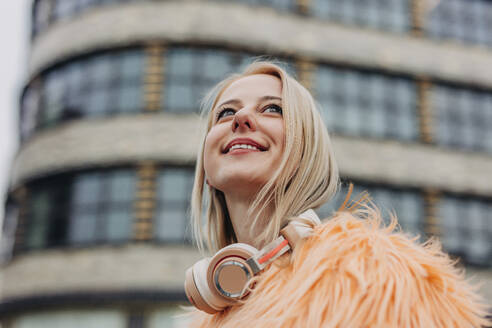 Lächelnde blonde Frau mit drahtlosen Kopfhörern vor einem Gebäude - VSNF01574
