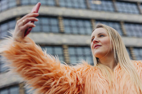 Frau trägt Pelzmantel und nimmt Selfie durch Smartphone in der Nähe von Gebäude - VSNF01575