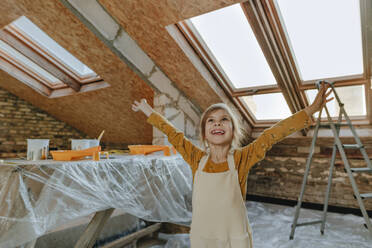 Cheerful girl standing with arms outstretched at house under construction - YTF01615