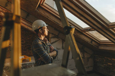 Architect wearing hardhat and looking out of window at construction site - YTF01620