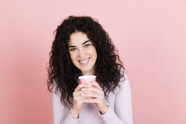 Happy woman holding disposable coffee cup against pink background - LMCF00799