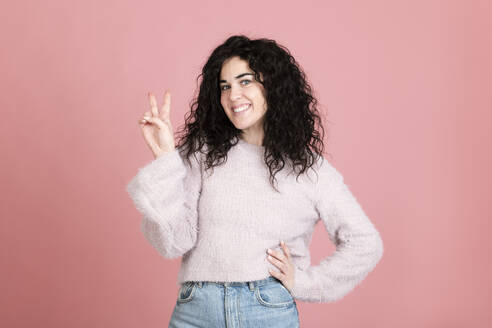 Happy young woman gesturing peace sign against pink background - LMCF00811