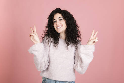 Smiling young woman gesturing peace sign against pink background - LMCF00813