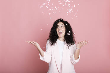 Cheerful young woman catching confetti against pink background - LMCF00828