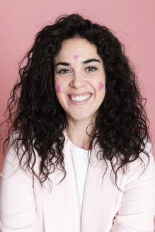 Glückliche Frau mit herzförmigen Aufklebern im Gesicht vor rosa Hintergrund - LMCF00832