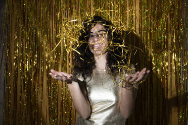 Happy woman playing with confetti in front of tinsel curtain - LMCF00841