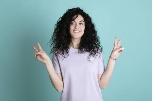 Smiling young woman gesturing peace sign against turquoise background - LMCF00842