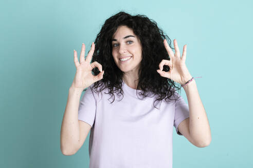 Smiling young woman gesturing OK sign against turquoise background - LMCF00843