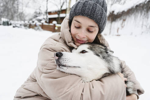 Lächelnde Frau umarmt Husky-Hund im Schnee - EYAF02930