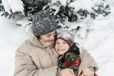 Glückliche Frau, die ihren Sohn umarmt und in der Nähe eines Baumes im Schnee liegt - EYAF02933
