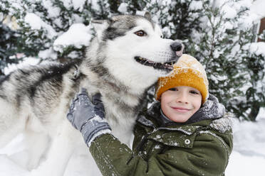 Lächelnder Junge streichelt Husky-Hund im Schnee in der Nähe eines Baumes - EYAF02934