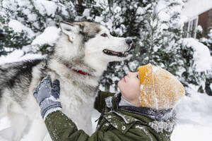 Boy petting husky dog near tree in snow - EYAF02935