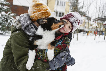 Verspielte Geschwister, die ihren Hund auf dem Schnee in den Armen halten - EYAF02937