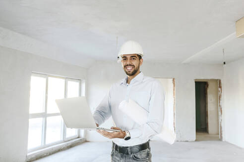 Happy architect standing with blueprint and laptop at site - AAZF01396