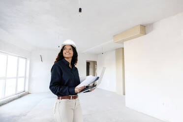 Smiling young architect standing with blueprint and laptop at site - AAZF01413