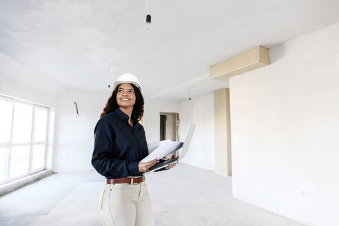 Lächelnder junger Architekt mit Bauplan und Laptop auf der Baustelle - AAZF01413