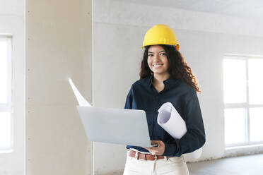 Lächelnder junger Architekt mit Laptop und Bauplänen auf der Baustelle - AAZF01417