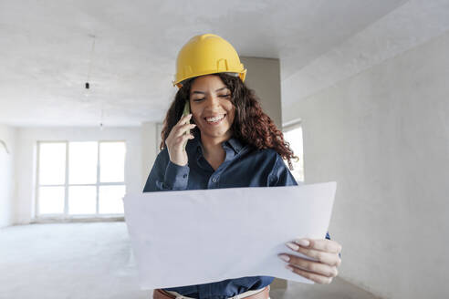 Zufriedener Architekt, der auf der Baustelle mit seinem Mobiltelefon über einen Bauplan diskutiert - AAZF01421