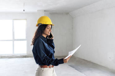 Smiling young architect having discussion over blueprint on mobile phone at site - AAZF01424