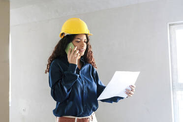 Young architect having discussion over blueprint on mobile phone at construction site - AAZF01425