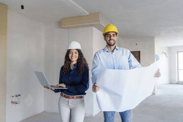 Smiling architects standing with laptop and blueprint at site - AAZF01433