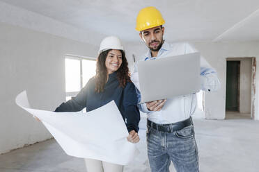 Smiling architect holding blueprint with colleague discussing over laptop at site - AAZF01439