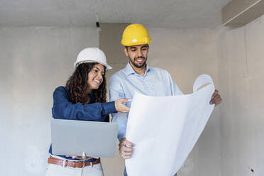 Happy architects discussing with blueprint and laptop at site - AAZF01441