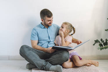 Glücklicher Vater liest seiner Tochter zu Hause vor der Wand ein Buch vor - OLRF00109