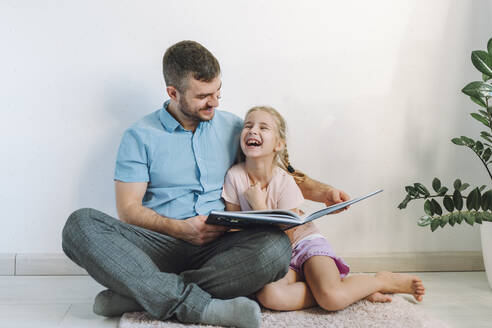 Happy man reading book to daughter in front of wall at home - OLRF00110