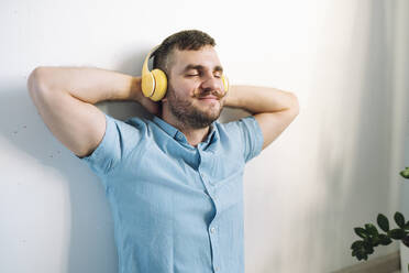 Man leaning on wall and listening to music through wireless headphones at home - OLRF00111