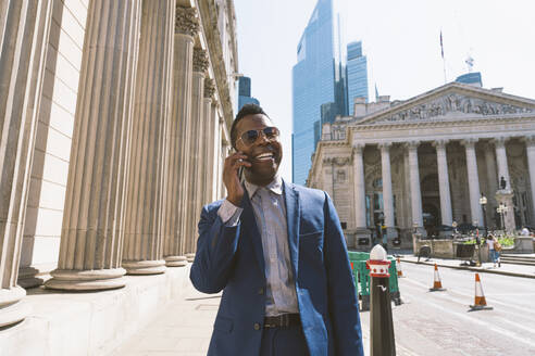 Lächelnder reifer Geschäftsmann, der in der Londoner Innenstadt mit einem Smartphone spricht - OIPF03772