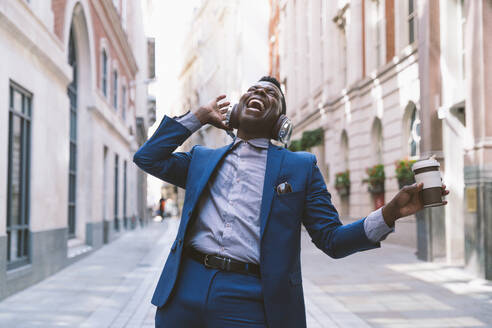 Cheerful mature businessman wearing wireless headphones holding coffee cup and enjoying on street - OIPF03784