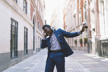 Happy mature businessman wearing wireless headphones holding coffee cup and dancing on street near buildings - OIPF03785