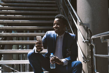 Lächelnder Geschäftsmann, der eine Wasserflasche hält und sein Smartphone auf einer Treppe benutzt - OIPF03798
