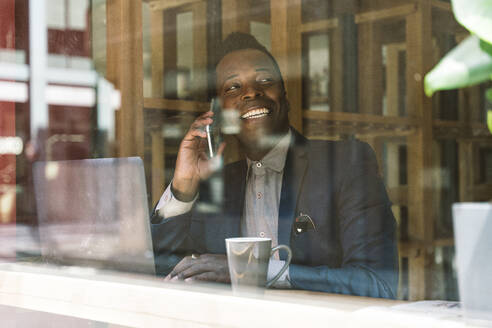 Glücklicher Geschäftsmann, der durch ein Glas im Café mit seinem Smartphone telefoniert - OIPF03810