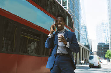 Lächelnder Geschäftsmann, der in der Nähe des Busses auf der Straße mit seinem Smartphone telefoniert - OIPF03817