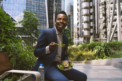 Happy businessman sitting with bowl of salad near buildings at office park - OIPF03822